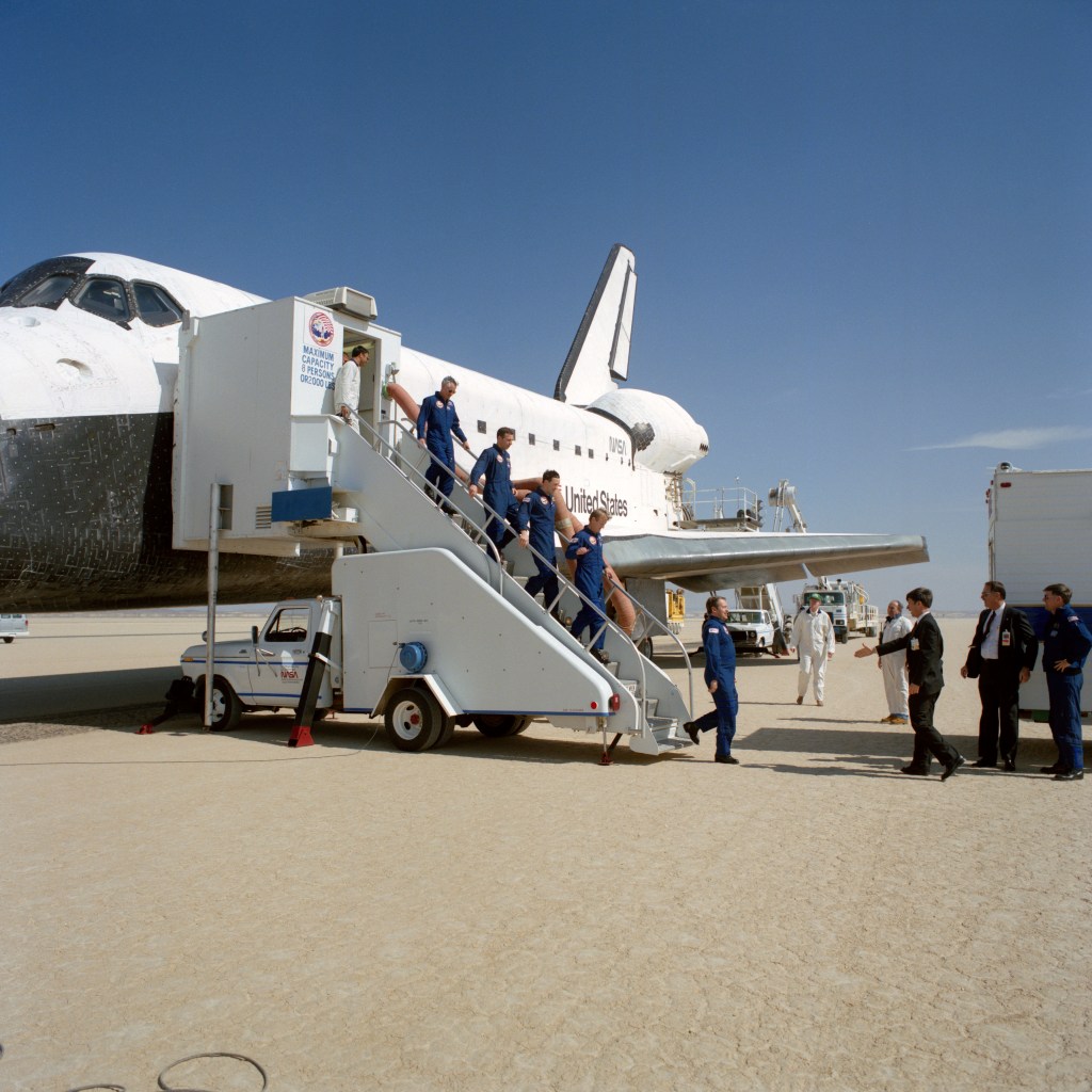 Five astronauts wearing blue flight suits descend the steps from the space shuttle onto the runway, greeted by officials.