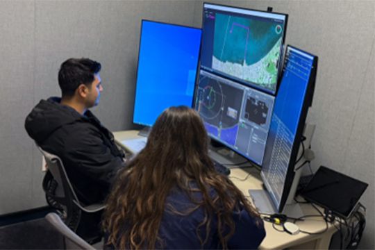 A researcher and a study participant sitting in front of the Ground Control Station in a HITL simulation to demonstrate how a set of NASA developed services, functions, and capabilities (SFCs) could be used in operational sUAS hurricane relief and recovery scenarios.