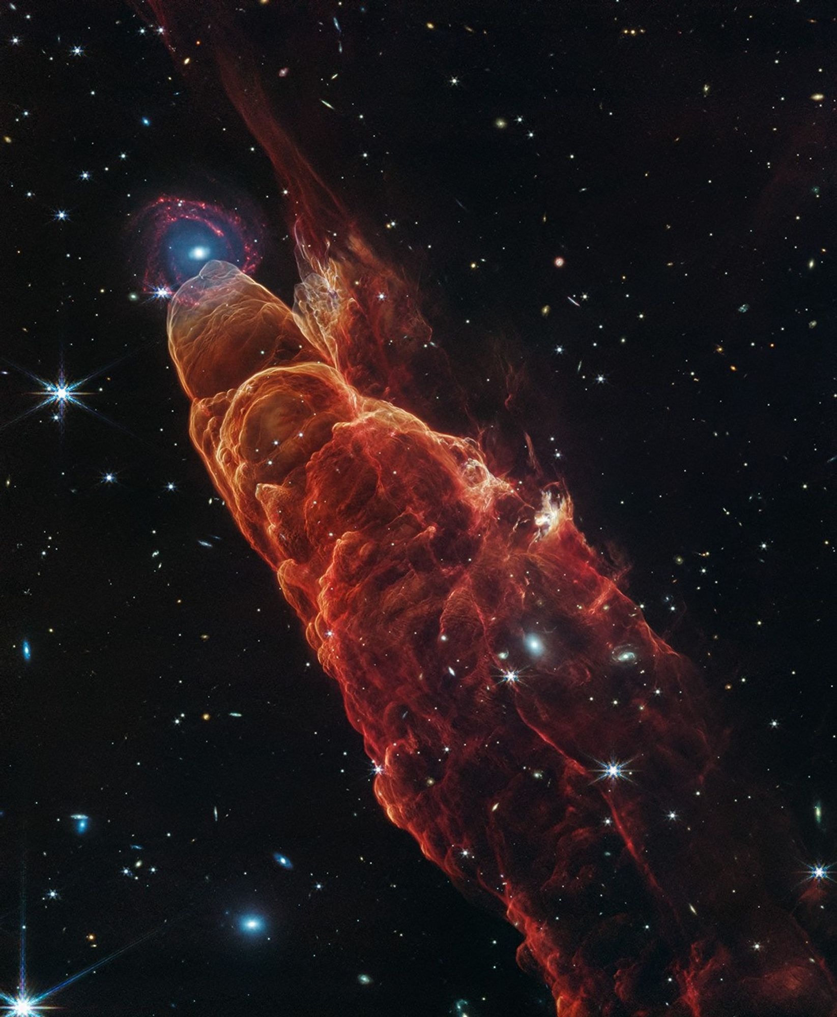 NASA’s James Webb Space Telescope observed Herbig-Haro 49/50, an outflow from a nearby still-forming star, in high-resolution near- and mid-infrared light. The intricate features of the outflow, represented in reddish-orange color, provide detailed clues about how young stars form and how their jet activity affects the environment around them. Like the wake of a speeding boat, the bow shocks in this image have an arc-like appearance as the fast-moving jet from the young star slams into the surrounding dust and gas. A chance alignment in this direction of the sky provides a beautiful juxtaposition of this nearby Herbig-Haro object with a more distant spiral galaxy in the background. Herbig-Haro 49/50 gives researchers insights into the early phases of the formation of low-mass stars similar to our own Sun. In this Webb image, blue represents light at 2.0-microns (F200W), cyan represents light at 3.3-microns (F335M), green is 4.4-microns (F444W), orange is 4.7-microns (F470N), and red is 7.7-microns (F770W).