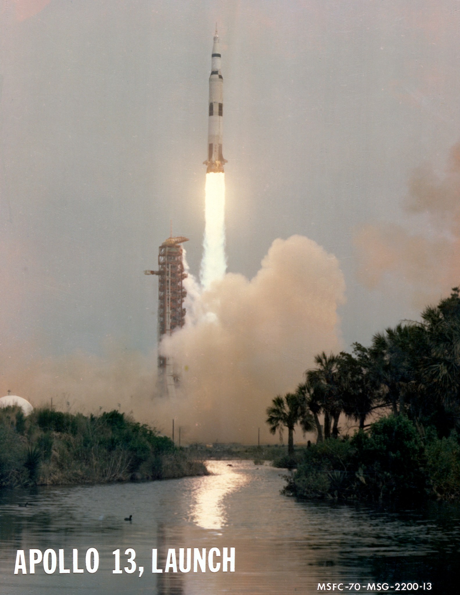 Apollo 13 launches upward, leaving a column of flames and light brown vapor behind it. Trees and vegetation frame the water at the bottom of the photo. Outlines of small animals can be see in the water. At bottom left is written "Apollo 13, launch" in capital letters. At bottom right is a series of letters and numbers labeling the photo.