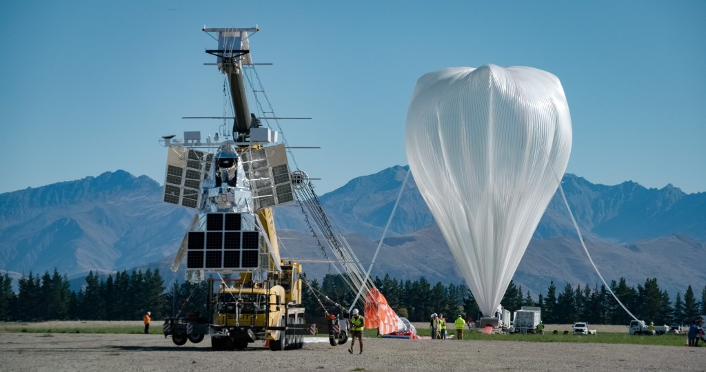 Balloon during inflation with New Zealand mountains in the background. The foreground depicts the SuperBIT balloon payload suspended by a crane.