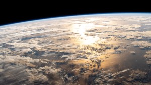 A view of Earth from the International Space Station. The Atlantic Ocean spreads out in front of us, with Earth's atmosphere appearing as a thin, hazy, light blue line bordering Earth. Sunlight shines off of the middle of the ocean, highlighting the edges of clouds over the water.