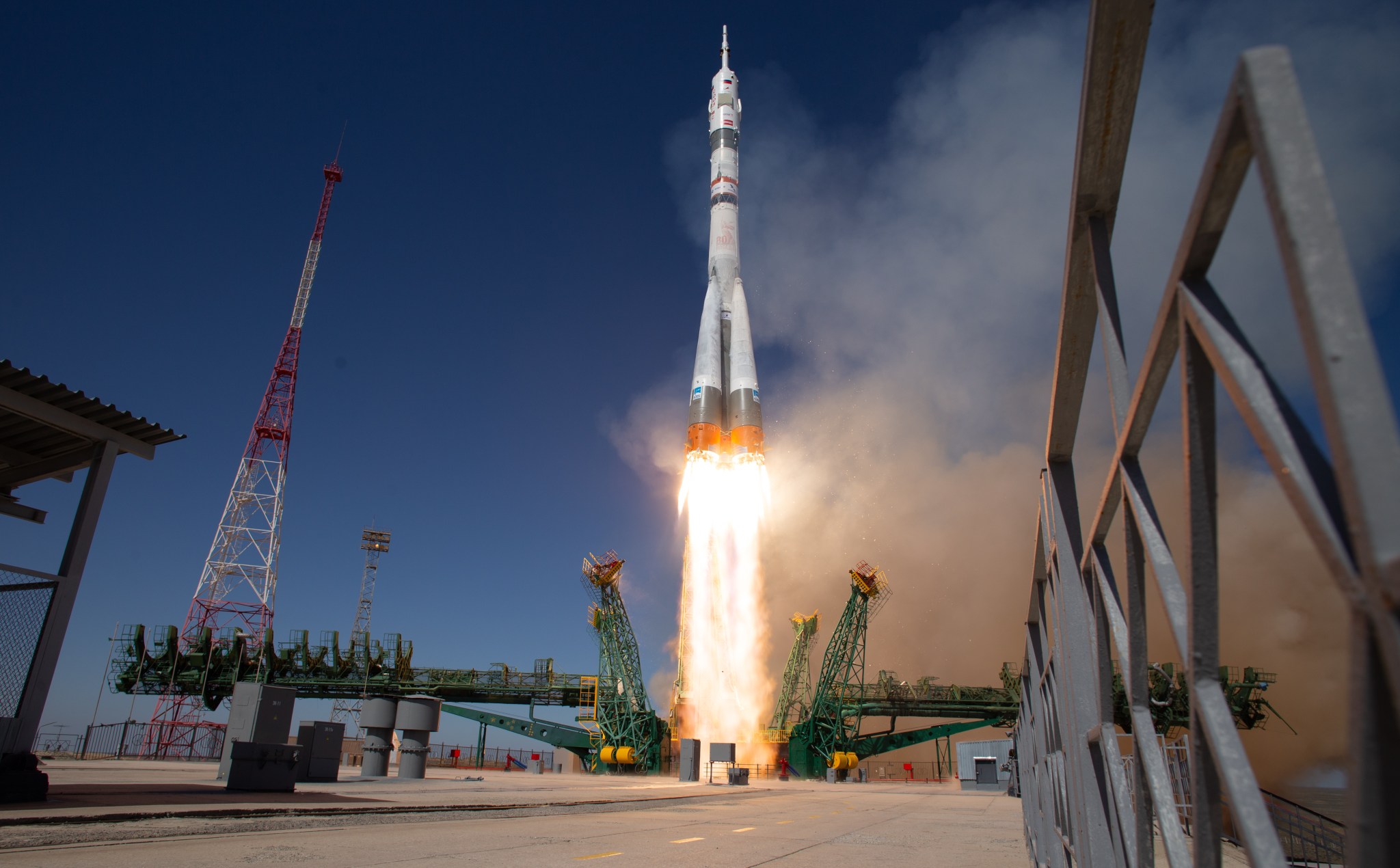 A light gray rocket lifts off, leaving flames and light brown vapor in its wake. Green supports lie perpendicular to the rocket.