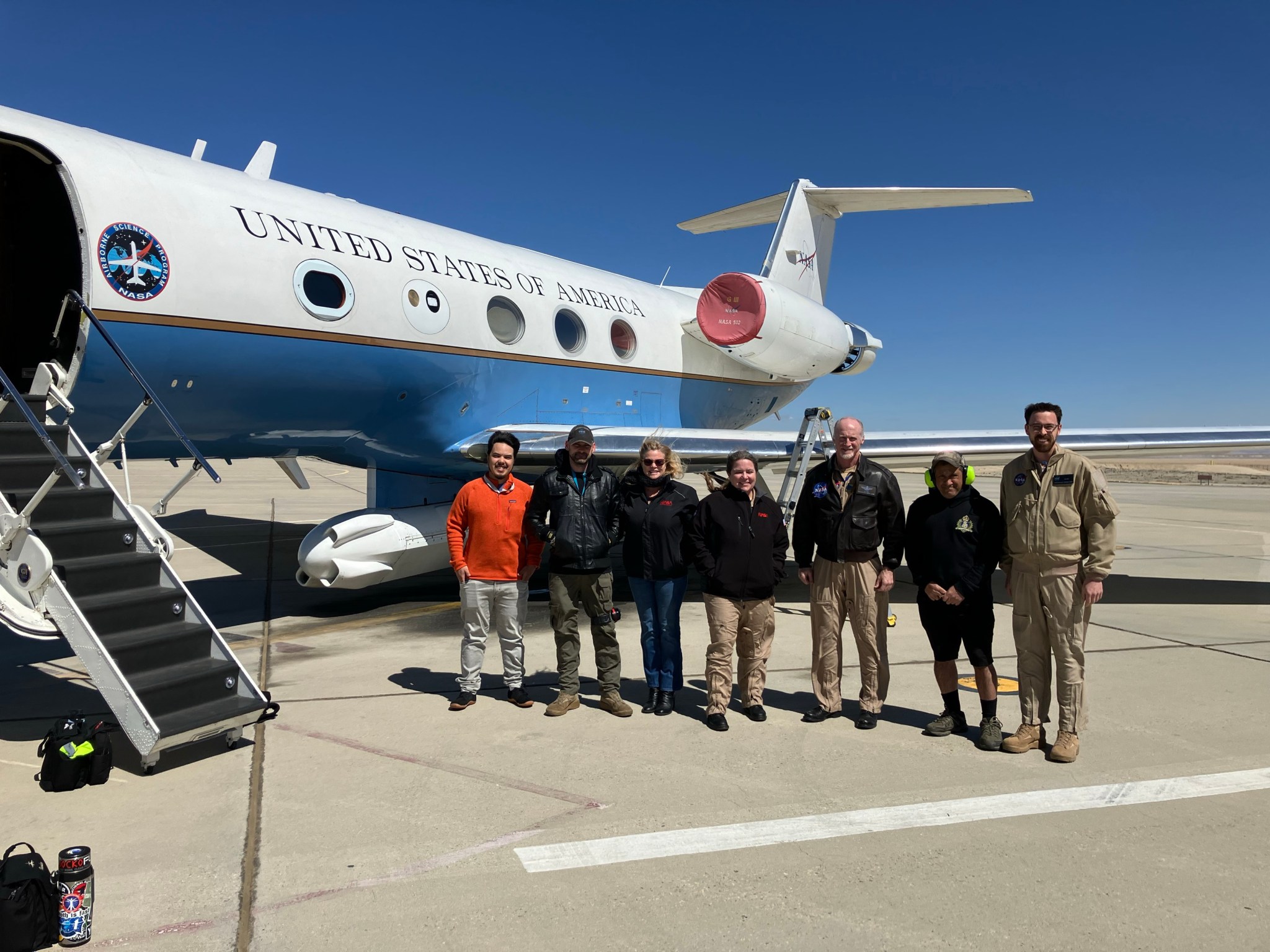Seven people stand next to an aircraft on a cement tarmac. They are posing for a team photo. All but the nose of the aircraft is visible in the left half of the frame, with its entry stairwell extended open. All team members are wearing jackets, and the wind appears to be blowing against them. Three of the team members appear to be wearing flight suits beneath their jackets, and one person is wearing plastic earmuffs for hearing protection.