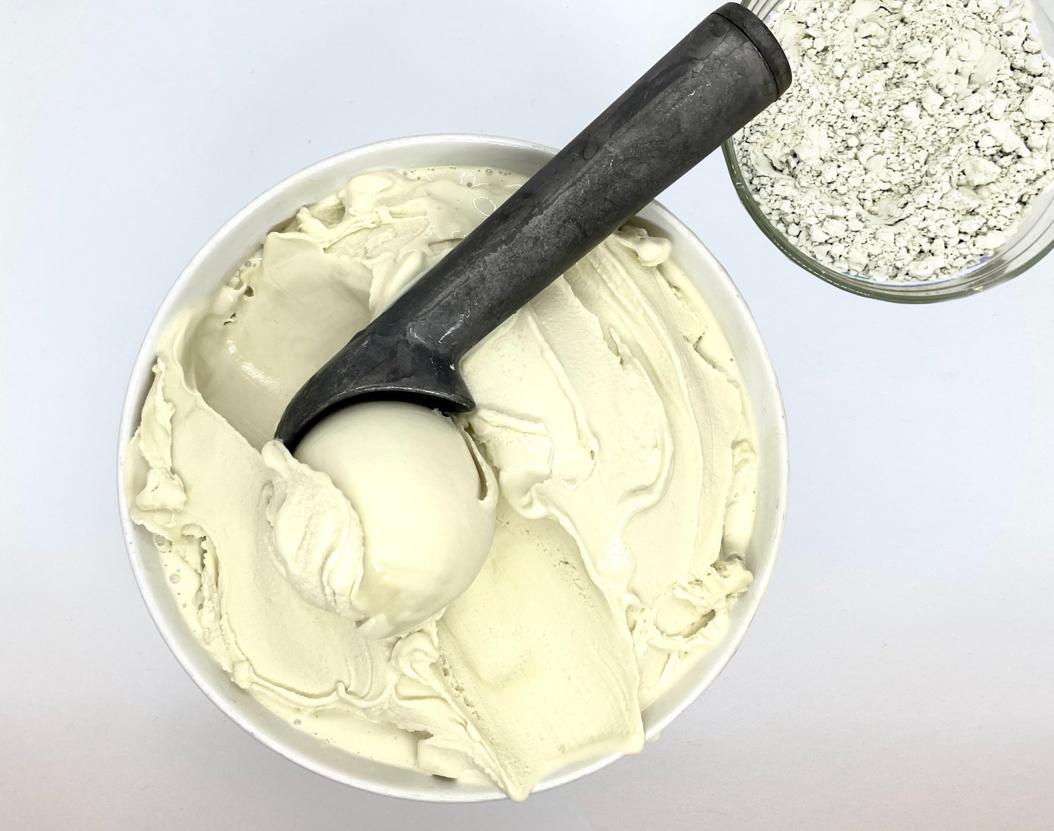 This image looks down on a white bowl filled with a creamy, slightly off-white substance and a metal ice cream scoop holding a ball of it. Next to the bowl is a smaller jar containing a coarse white powder.