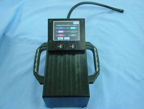 A black handheld device with a digital screen displays combustion product levels, including carbon dioxide and carbon monoxide. It has two switches and a hose attached and rests on a light blue surface.