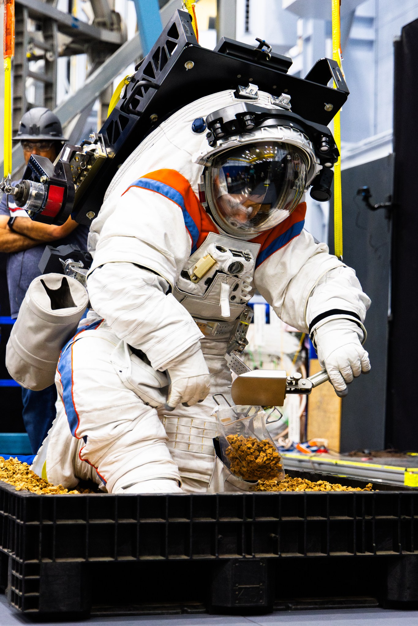 An engineer in a white spacesuit with red and blue accents kneels in a sandbox, using a scoop to collect simulated lunar soil into a bag during a training or equipment test indoors. A harness supports the suit from above.