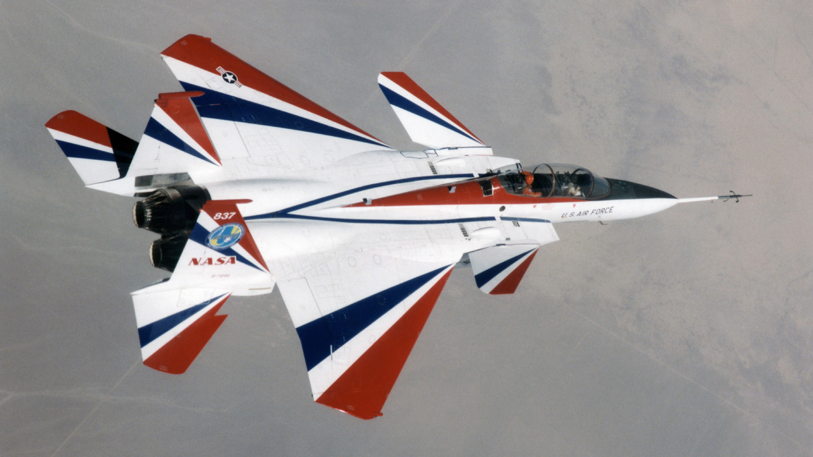 A NASA and U.S. Air Force jet with red, white, and blue markings flies over a desert landscape, viewed from above. The aircraft features sleek lines and a sharp nose, with visible NASA and military insignias.
