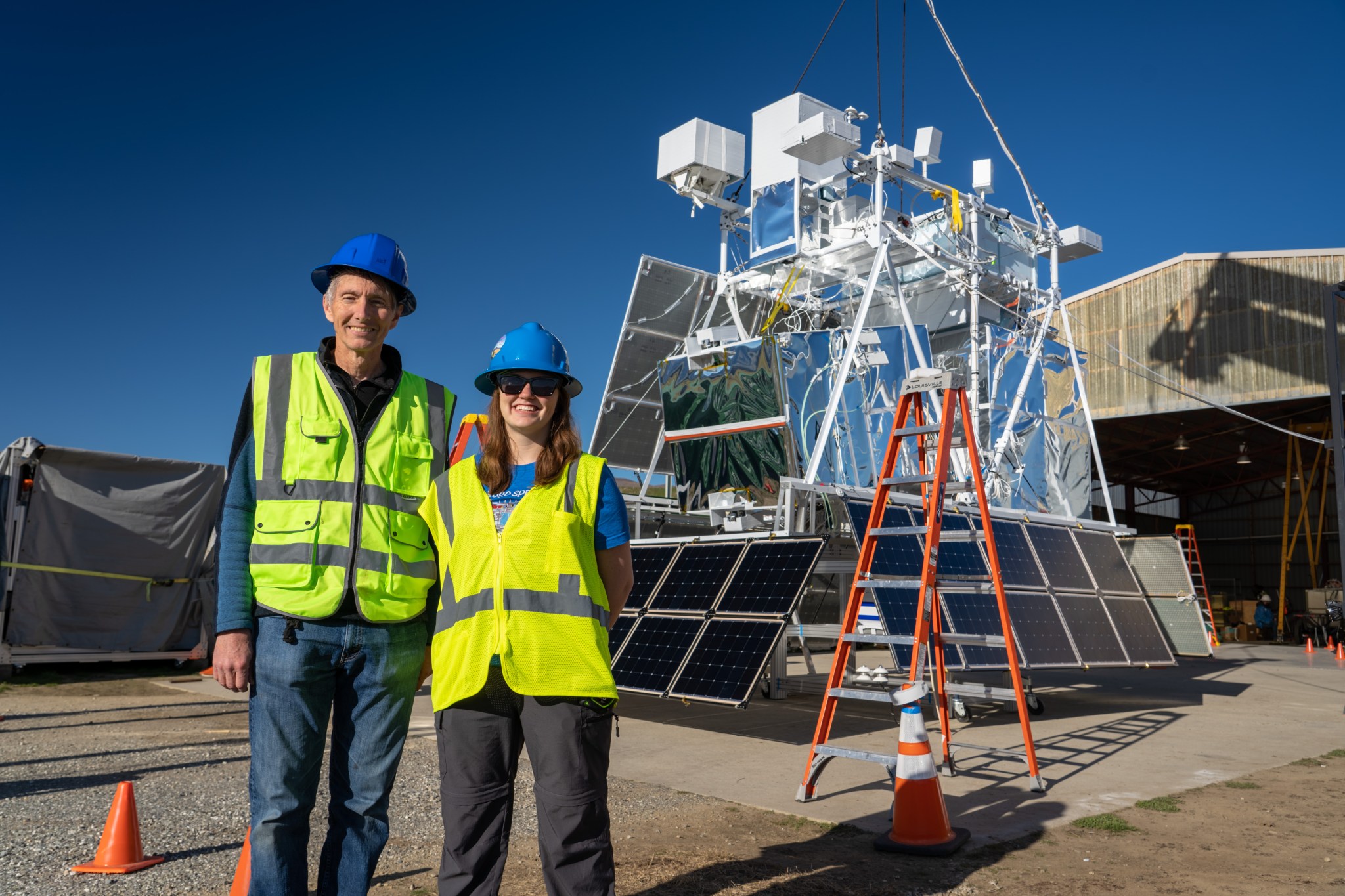 two people stand infront of a science payload wearing bright yellow safety vests