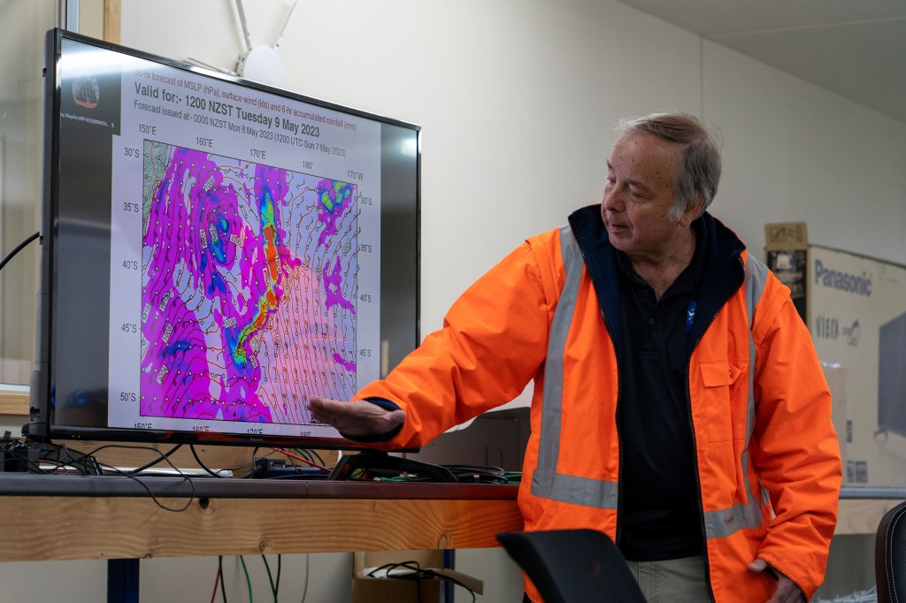 Meteorologist in a safety orange jacket, gestures toward a wind map displayed on a TV screen