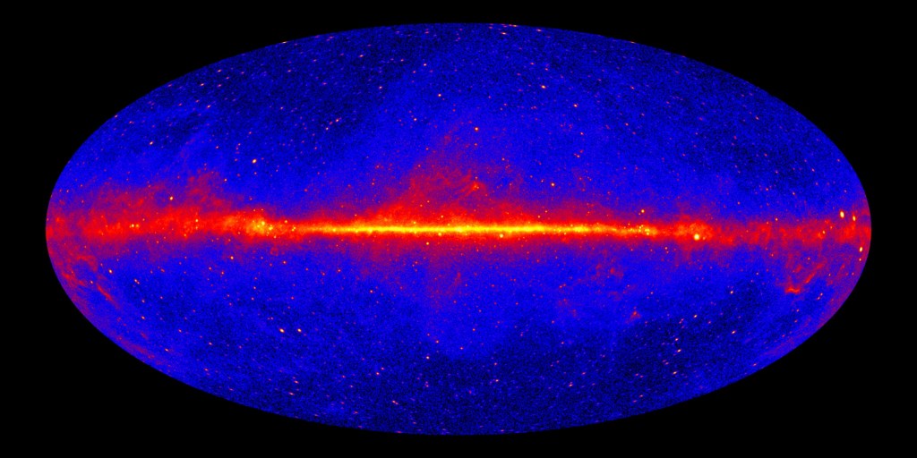 A data visualization of gamma ray observations from the Fermi Gamma-ray Space Telescope. Brighter colors represent more intense sources. The visualization features a blue, horizontal oval framed by a black background. Its edges are darker due to large groups of dense black dots. These groups become sparse near the oval’s center, revealing a brighter blue color. A glowing yellow line emitting wispy red clouds spans horizontally across the oval’s center. Small, bright orange dots are scattered across the shape’s entirety.