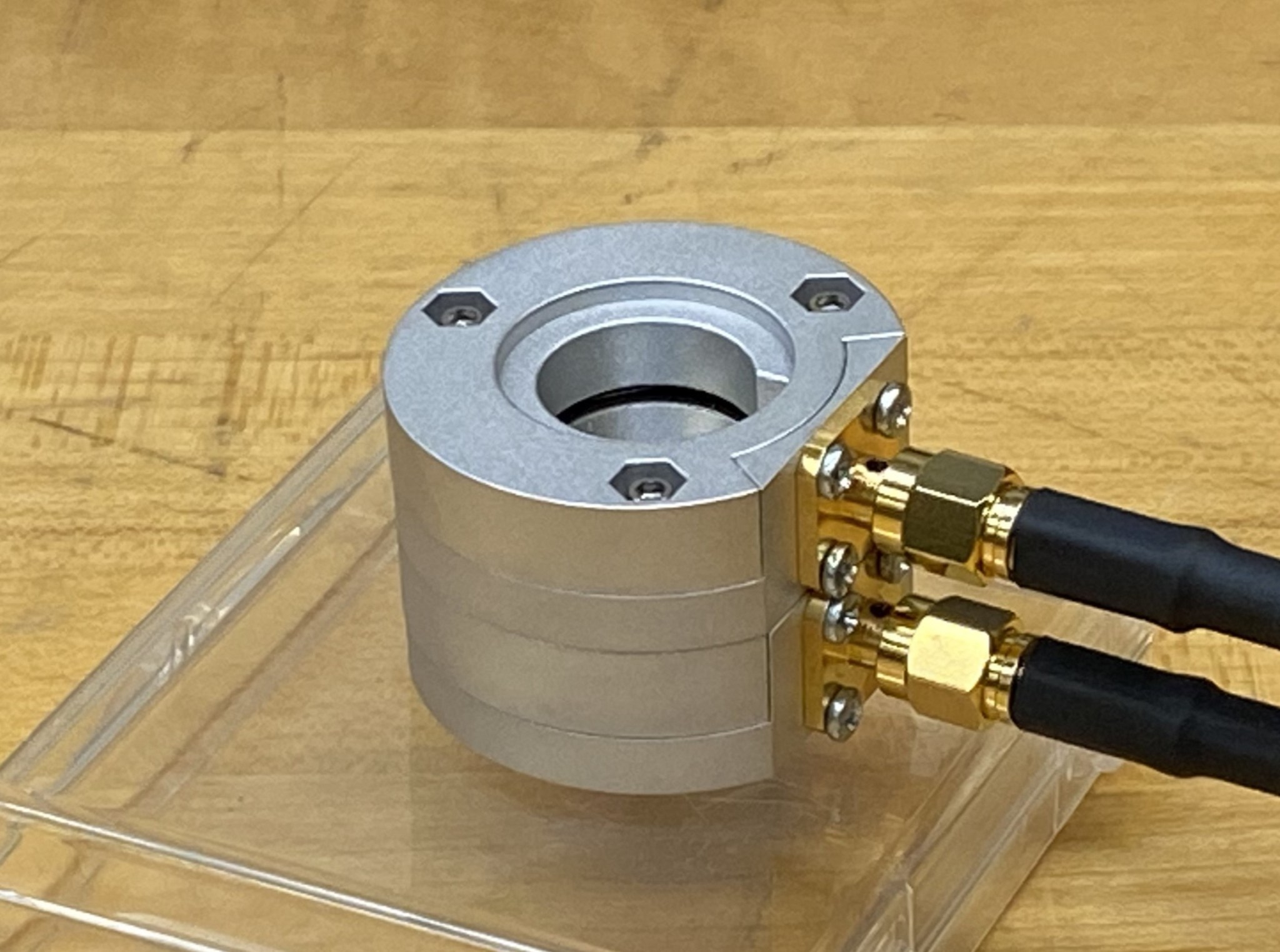 Metal cylinder with a flat semiconductor detector visible through an opening in the top with two cables attached to the right, set on a clear plastic box on a wood bench.