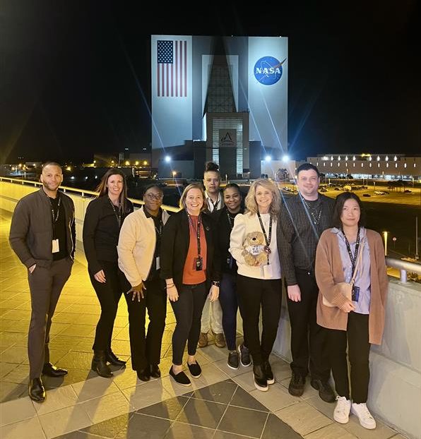 Nine people stand on a rooftop in front of a large building featuring the NASA meatball logo (right) and a U.S. flag.