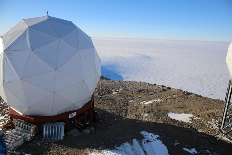 A large, white geodesic dome protecting a ground station antenna sits on a raised platform labeled KSAT at the Troll Satellite Station in Antarctica. The rocky, snow-dusted terrain sits in the foreground of an expanse of ice and a clear blue sky. A small group of people is visible at a distance near the edge of the plateau.