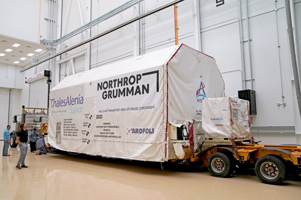The HALO transport container, wrapped in white material with logos for Artemis, Northrop Grumman, Thales Alenia Space, and other partners, is positioned inside Northrop Grumman’s facility in Gilbert, Arizona. The container remains on a flatbed trailer as personnel monitor its arrival in the clean, white-walled space.