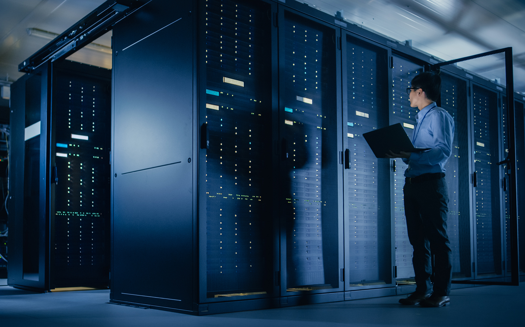 In a data center, a male IT Technician stands and looks at a laptop next to a server rack.