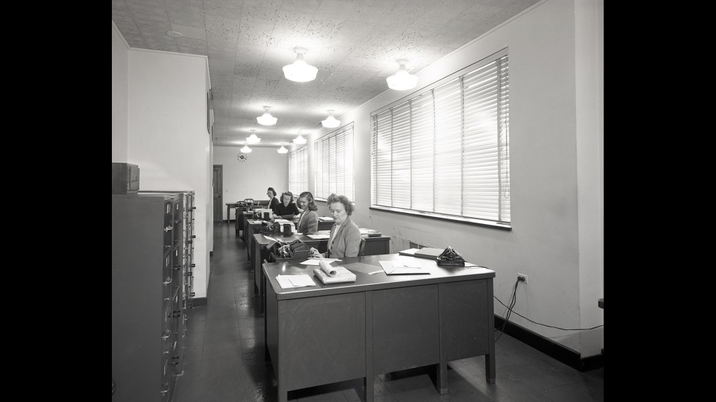 Women at desks in narrow room.