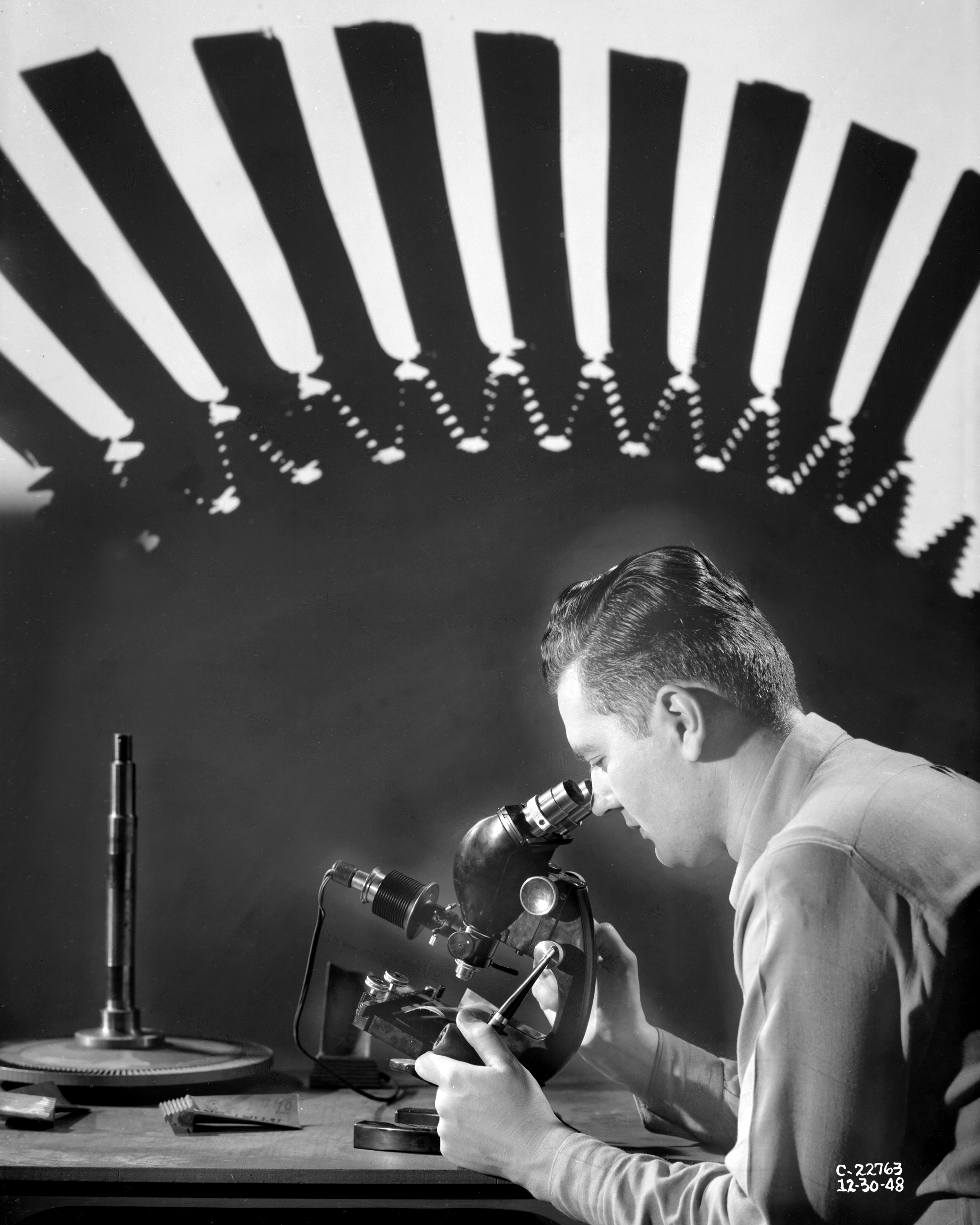 Composite of man looking into microscope with large turbine wheel in background.