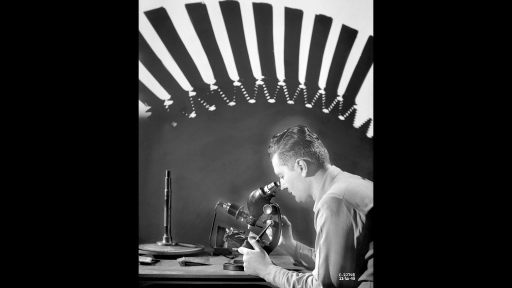 Composite of man looking into microscope with large turbine wheel in background.