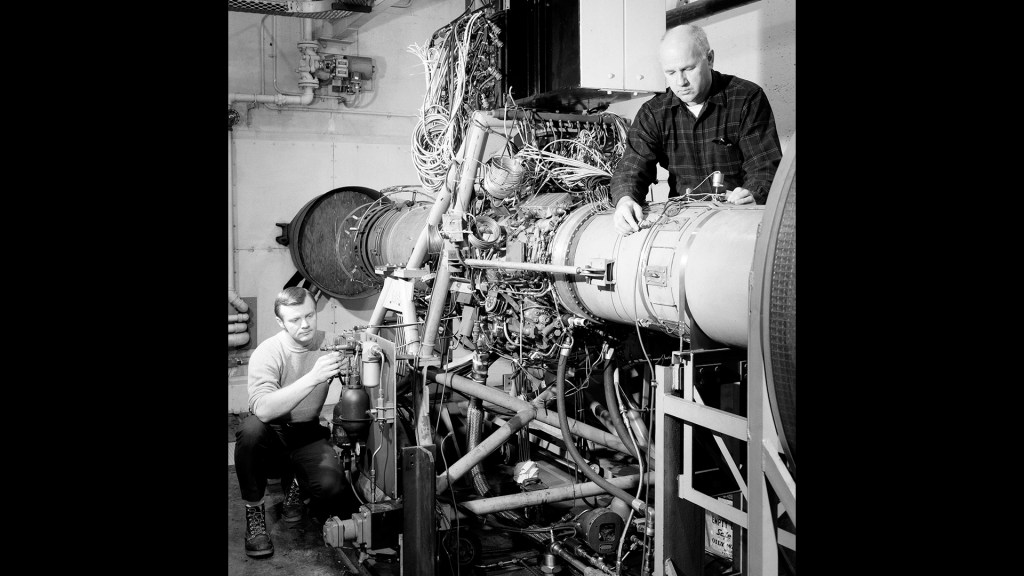 Two men instrumenting jet engine in test cell.