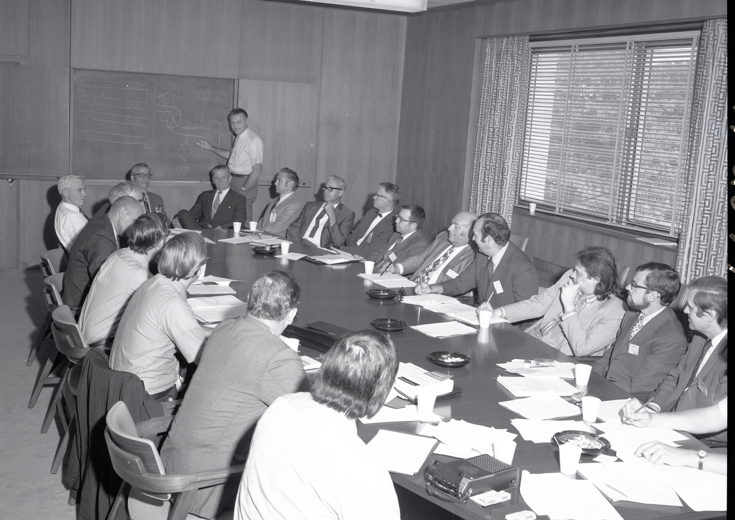Man at chalkboard and others sitting at conference table.