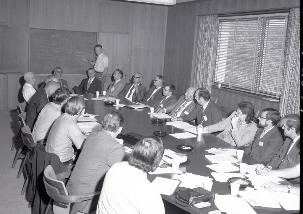 Man at chalkboard and others sitting at conference table.