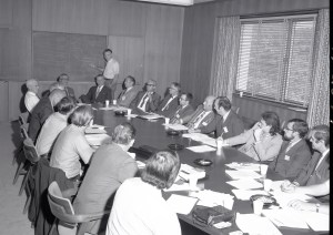 Man at chalkboard and others sitting at conference table.