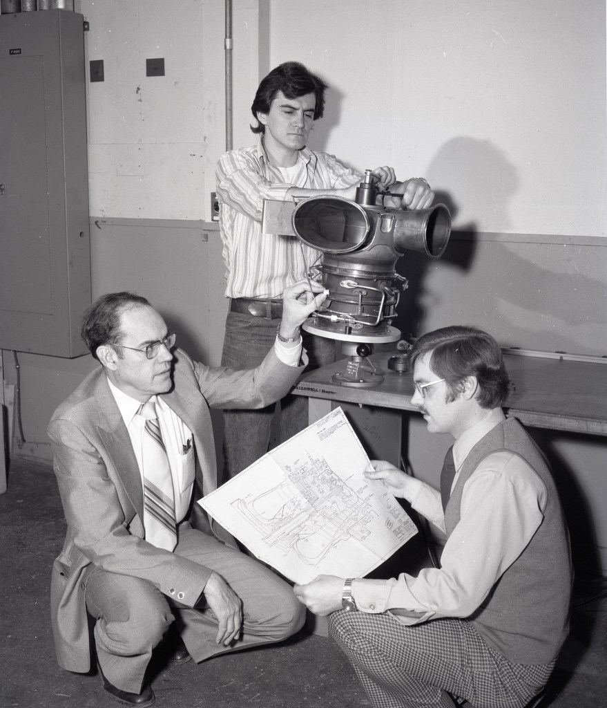 Three men looking at engine and blueprints.