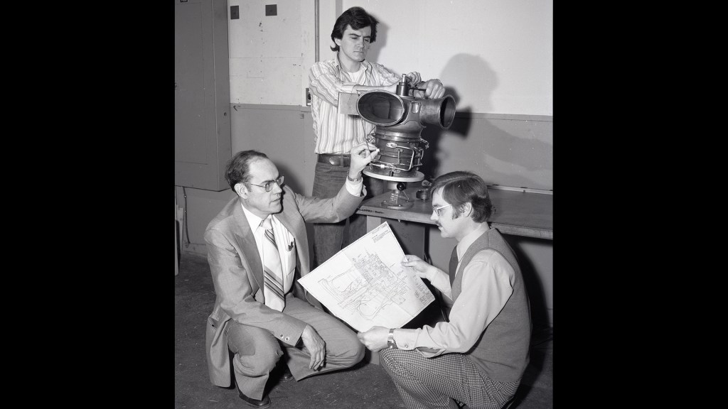 Three men looking at engine and blueprints.