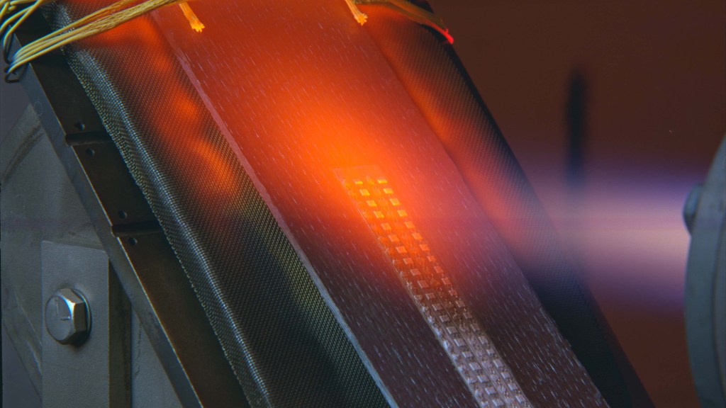 Close-up of ceramic matrix composite panel being tested with intense flames. The panel glows as it is exposed to the high temperature, with visible wiring at the bottom held in place with white cement. The material’s texture is visible, showcasing its heat-resistant properties.