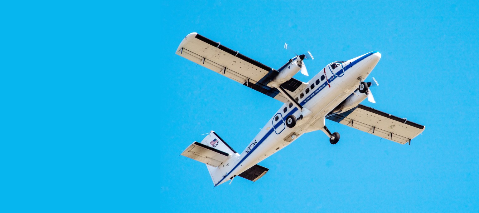 A twin-engine propeller-driven aircraft with a high wing is seen from below looking up with clear skies.