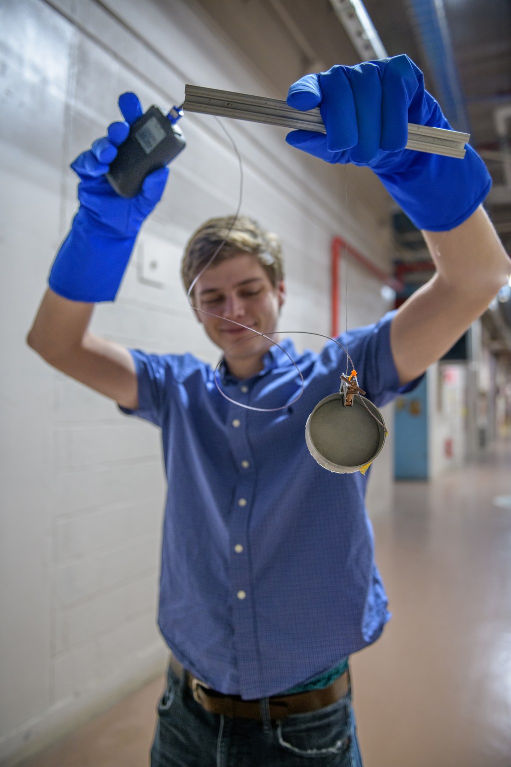 A man wearing blue rubber gloves and a blue button down shirt holds up a circular object attached to thin wires. 