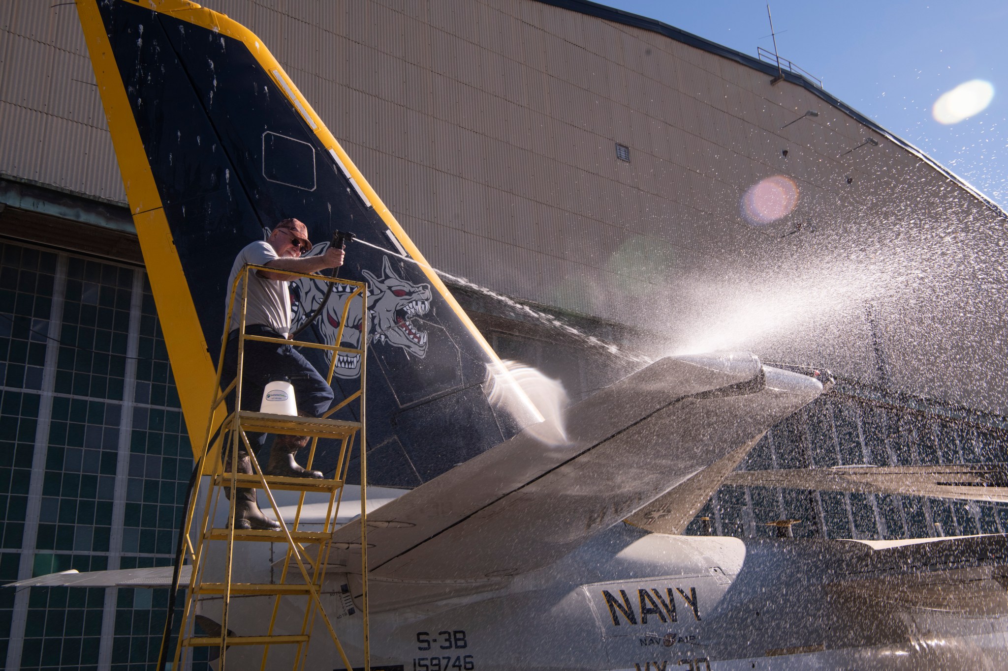 A man washes the S-3B Viking’s wings with a hose. He stands on a yellow ladder to reach the top of the aircraft’s wing. He wears sunglasses and a red hat. It is a sunny day, and sunlight reflects off the water from the hose. The aircraft is parked in front of the NASA Glenn Hangar. The wings are folded atop the aircraft.