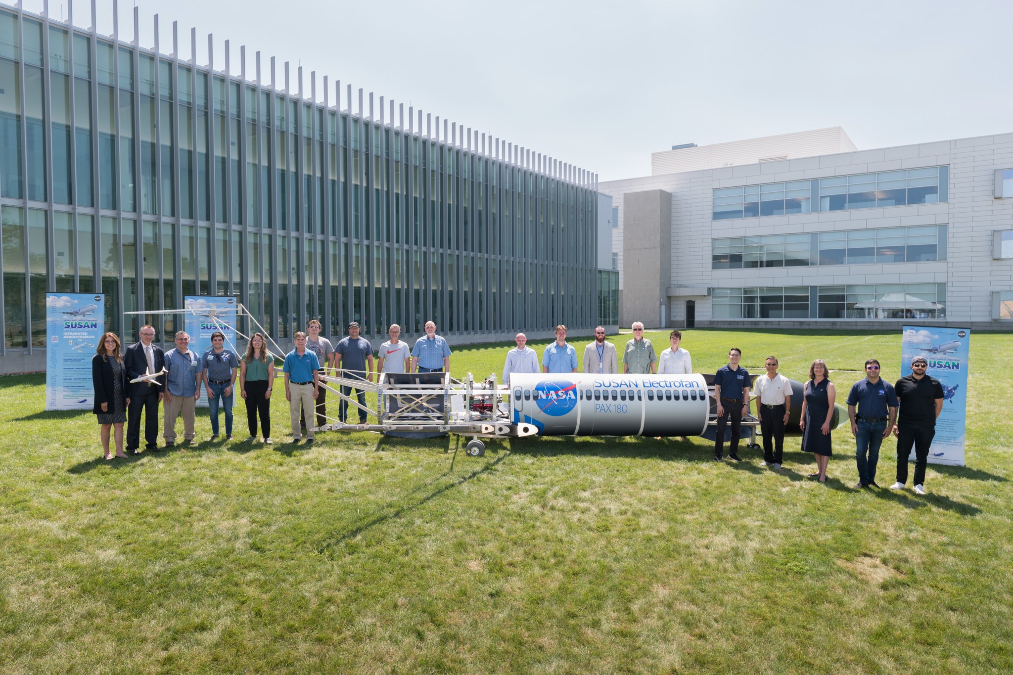 A group of people stand behind a model aircraft that sits on green grass in front of a large, gray building.