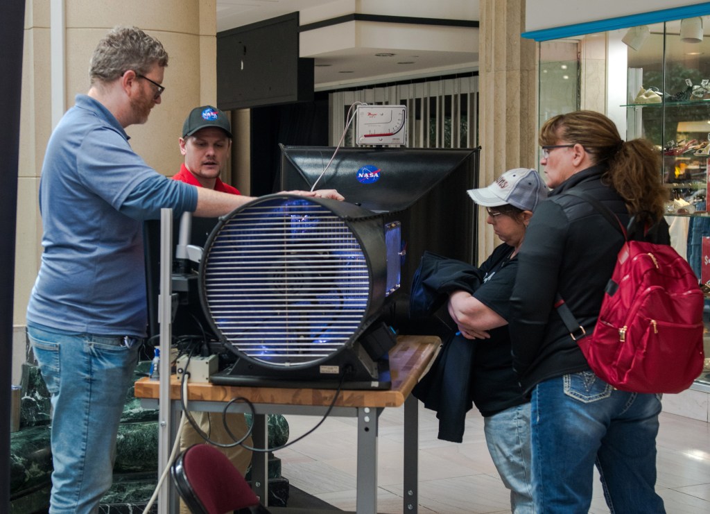 Two NASA Glenn employees stand to the left of a cylindrical, portable wind tunnel on a table and simulate a test ito showcase its capabilities. Two guests stand on the other side of the tunnel and observe the test.