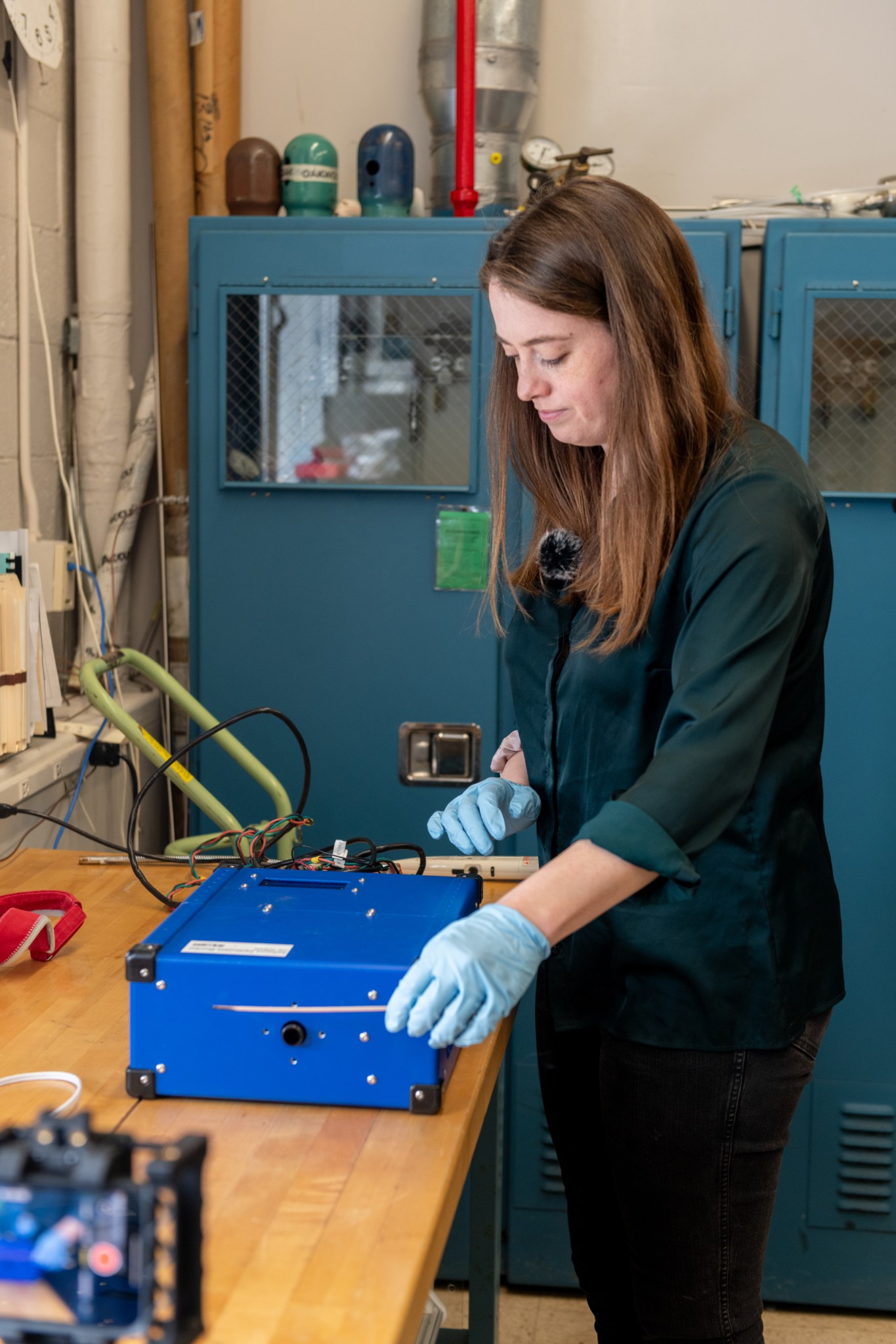 A woman scientist with auburn hair and emerald green button-down shirt holds stands next to a blue air quality monitor and holds burning incense near it. She wears blue gloves, and the monitor rests on a wooden table.