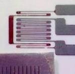 Close-up of an etched strain gauge sensor on a white surface. The sensor features a series of parallel metallic lines, forming a rectangular shape with connected tabs extending outward. A textured, dark rectangular reference ruler is in the bottom left.