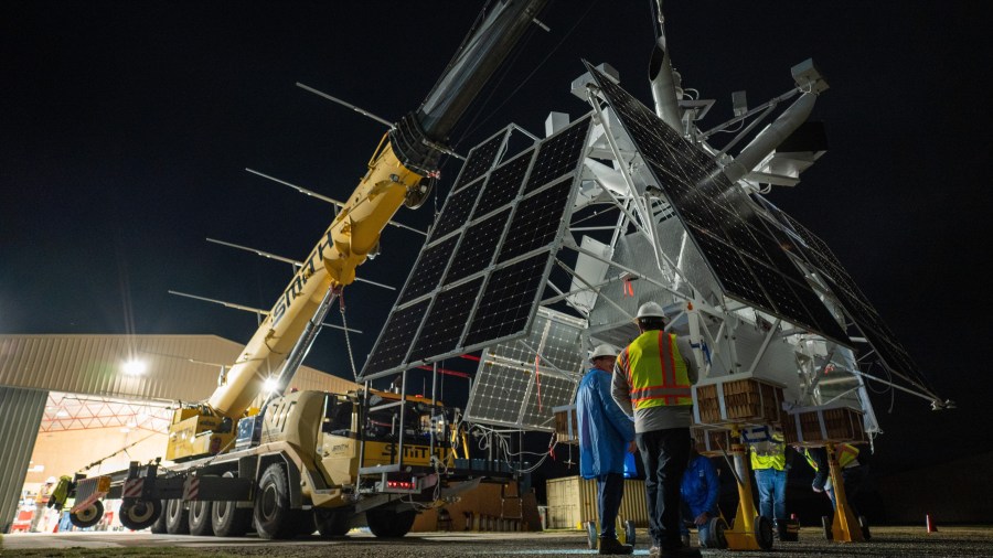 A large metal scientific instrument with black panels is suspended off the ground by a yellow crane.