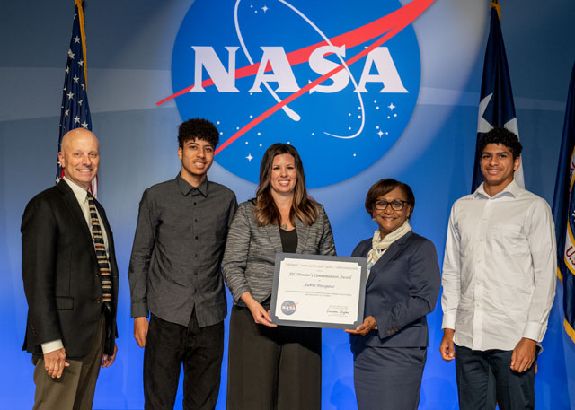 A group of five people stand in front of two flags and a NASA emblem. The woman in the middle holds an award.