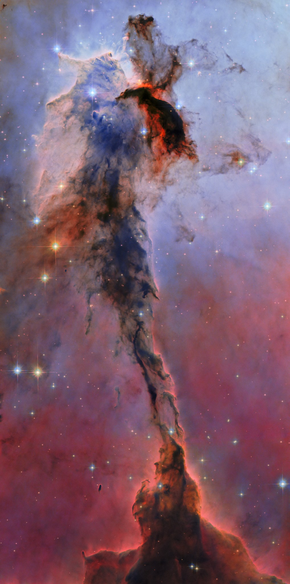 A tall, thin structure of dark gas clouds. This pillar is darker and broader at its base, thins toward the middle, and broadens out again at the top, with spikes, fingers, and wisps of gas protruding in all directions from its head. Some parts of the pillar are illuminated, but most are dark and silhouetted at the edges and lit from behind. A wall of colorful gas lies behind the pillar, bluish at the top and redder toward the bottom, with several blue and gold stars scattered across it.
