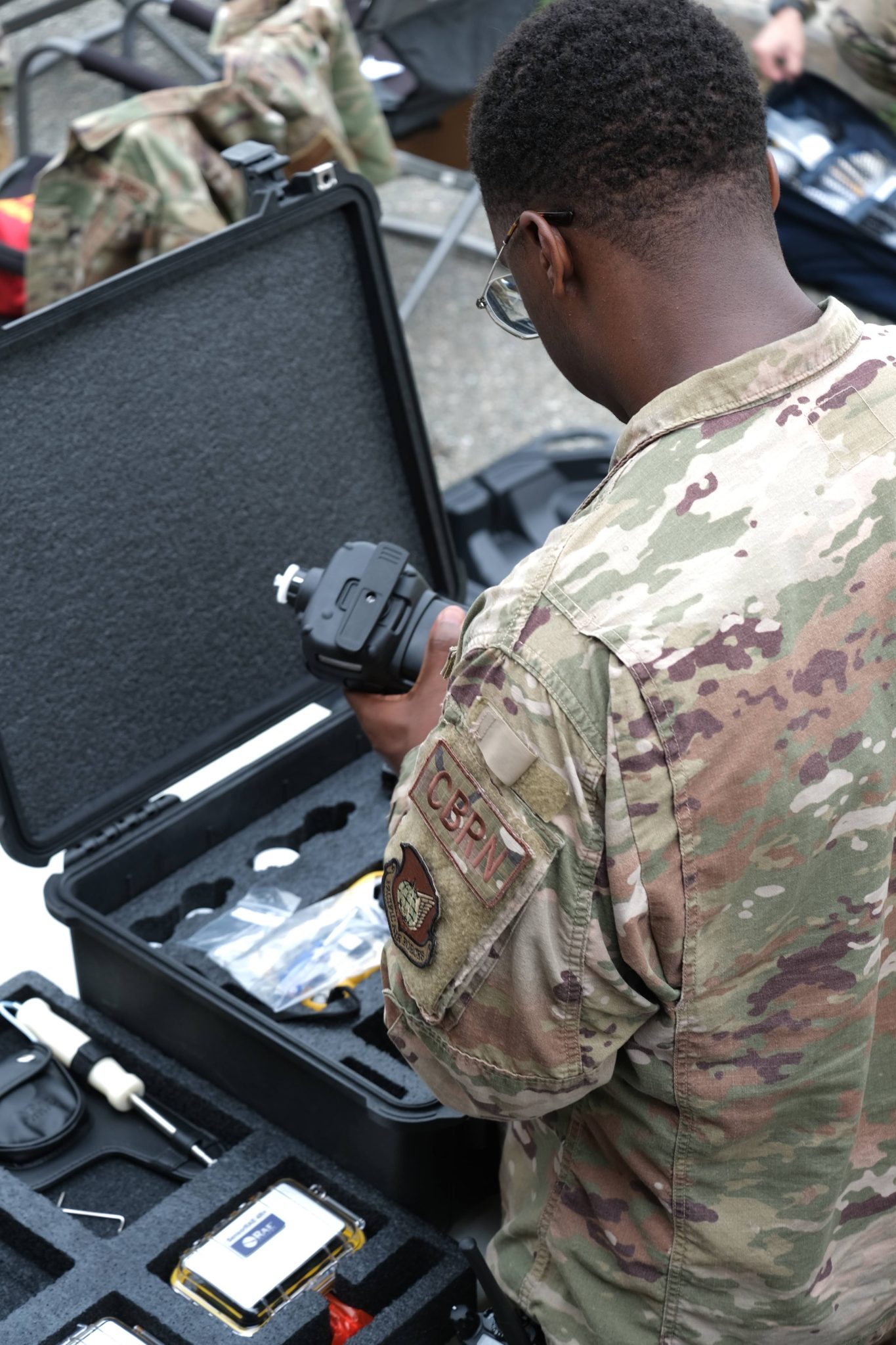 A U.S. military service member wearing camouflage uniform inspects a device from a protective foam case containing various technical equipment and tools.