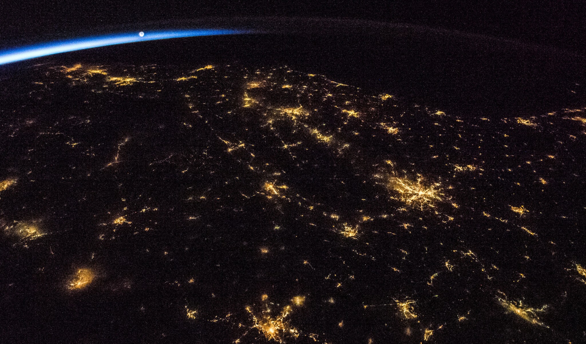 A nighttime view of Earth from space shows clusters of city lights scattered across the landscape, with a thin blue line of the atmosphere and a distant sunrise visible on the horizon.