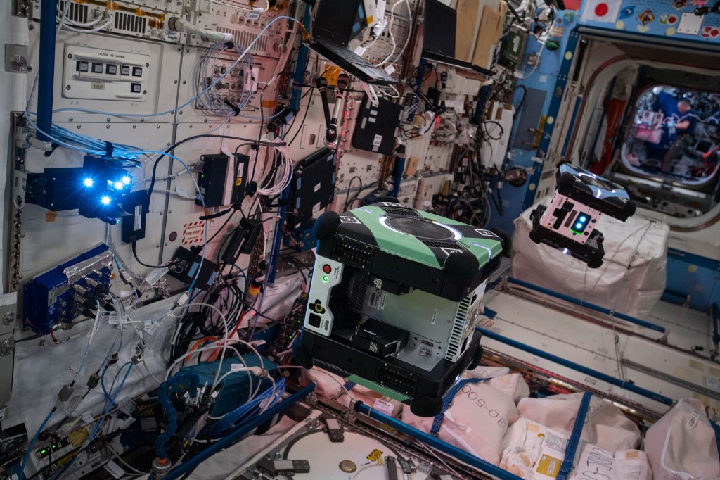 Two cube-shaped robots each about the size of a small suitcase, one green and one blue, float in the middle of a module on the space station. Below them are white storage bags and behind them a wall covered with equipment, laptops, cords, and wires. A black device mounted on the wall at the left of the image has four bright blue lights.