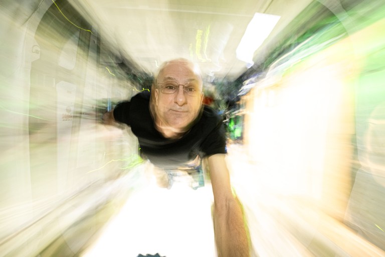 NASA astronaut Don Pettit takes an out-of-focus selfie facing the camera while floating through an International Space Station corridor. He is wearing glasses and a black t-shirt. The walls surrounding him are blurry and light yellow in color.