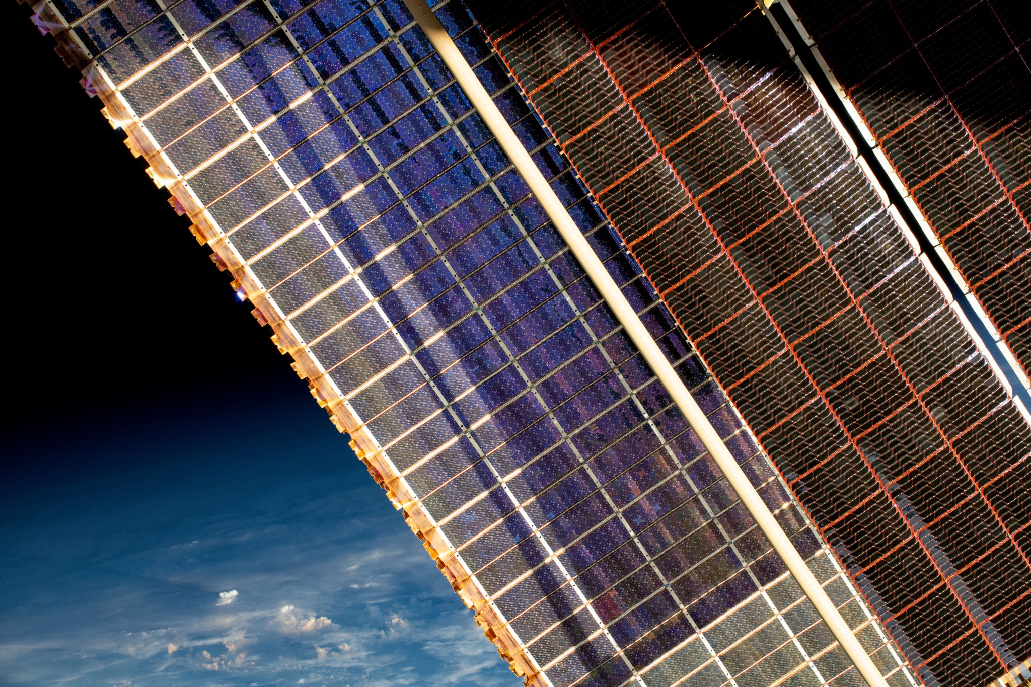 Two solar arrays fill this image diagonally. The solar array on the left has a rectangular grid pattern and is dark blue with a light brown outer edge. The solar array to the right is black with a red grid pattern and many parallel white lines within the grid. In the background is part of the Earth is blue, and space is black.