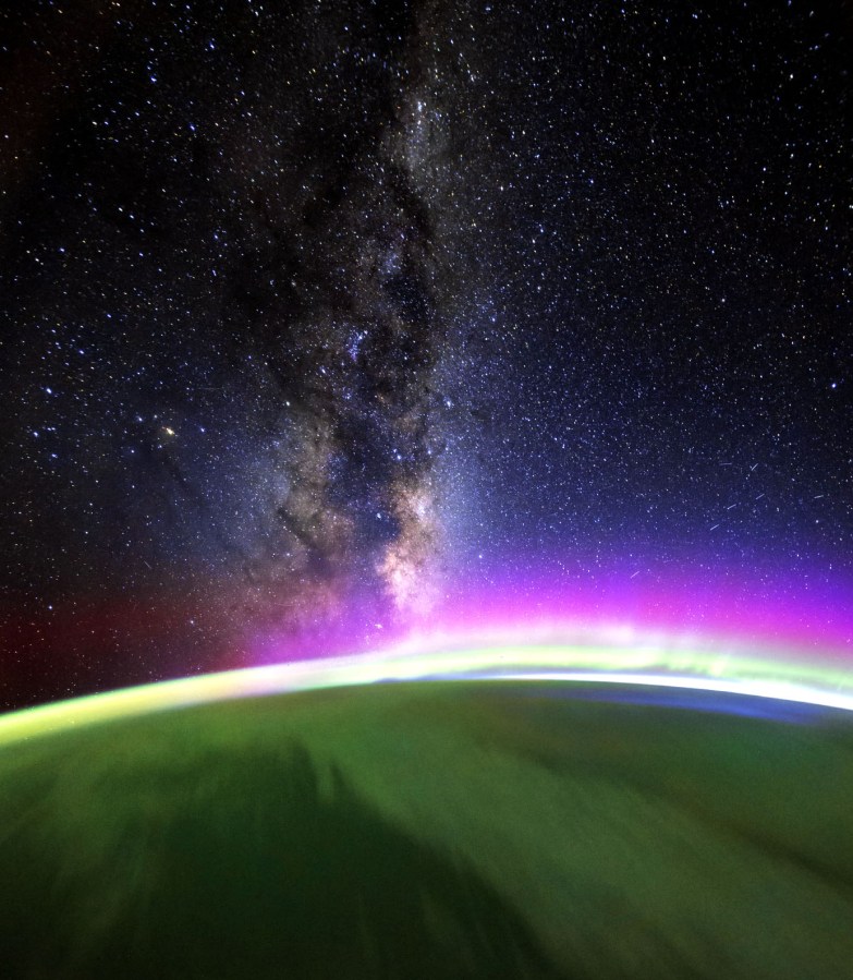 A small part of Earth from space is shown in dark green, and the edge of the curvature is illuminated with light that diffuses into space in shades of violet and then dark blue. In the background, a portion of the Milky Way runs perpendicular to the Earth’s curvature. Many other stars dot the area surrounding the Milky Way.