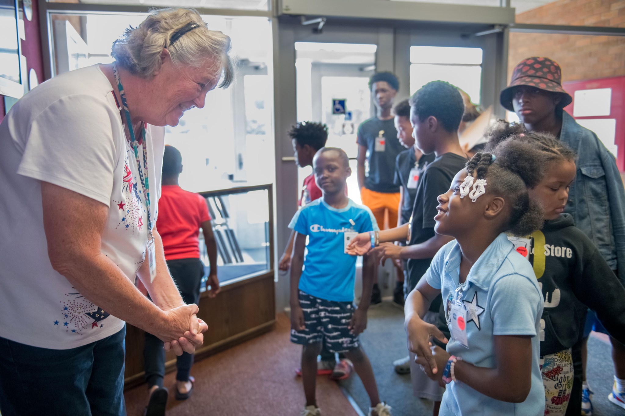 A group of grade school students explore the lobby of NASA Glenn's Telecscience Support Center, a facility where researchers operate International Space Station experiments. A NASA expert wearing a white shirt with blue and red stars and jeans clasps her hands and smiles at a student looking up at her. The student is a grade school girl with a light blue shirt and braids with white hair clips. She smiles up at the expert and appears to be asking a question. Other students look on in the background.