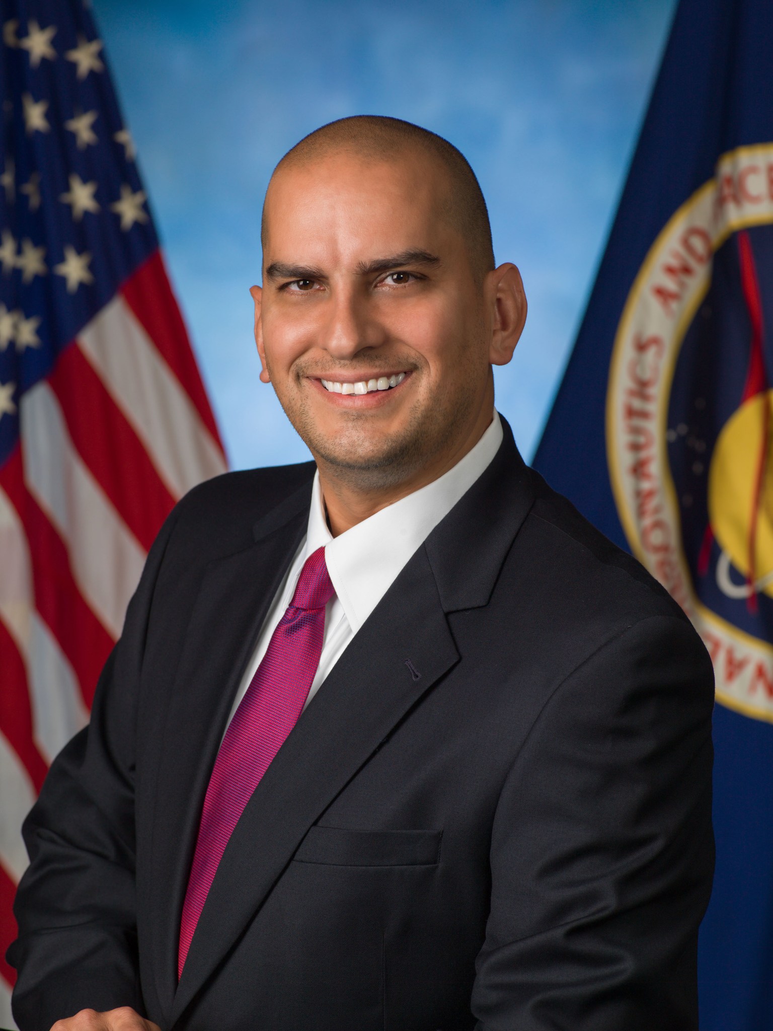 Official portrait of Johnson Space Center employee Jason Phillips, who wears a black suit, fuchsia tie, and stands in front of the NASA and U.S. flags.