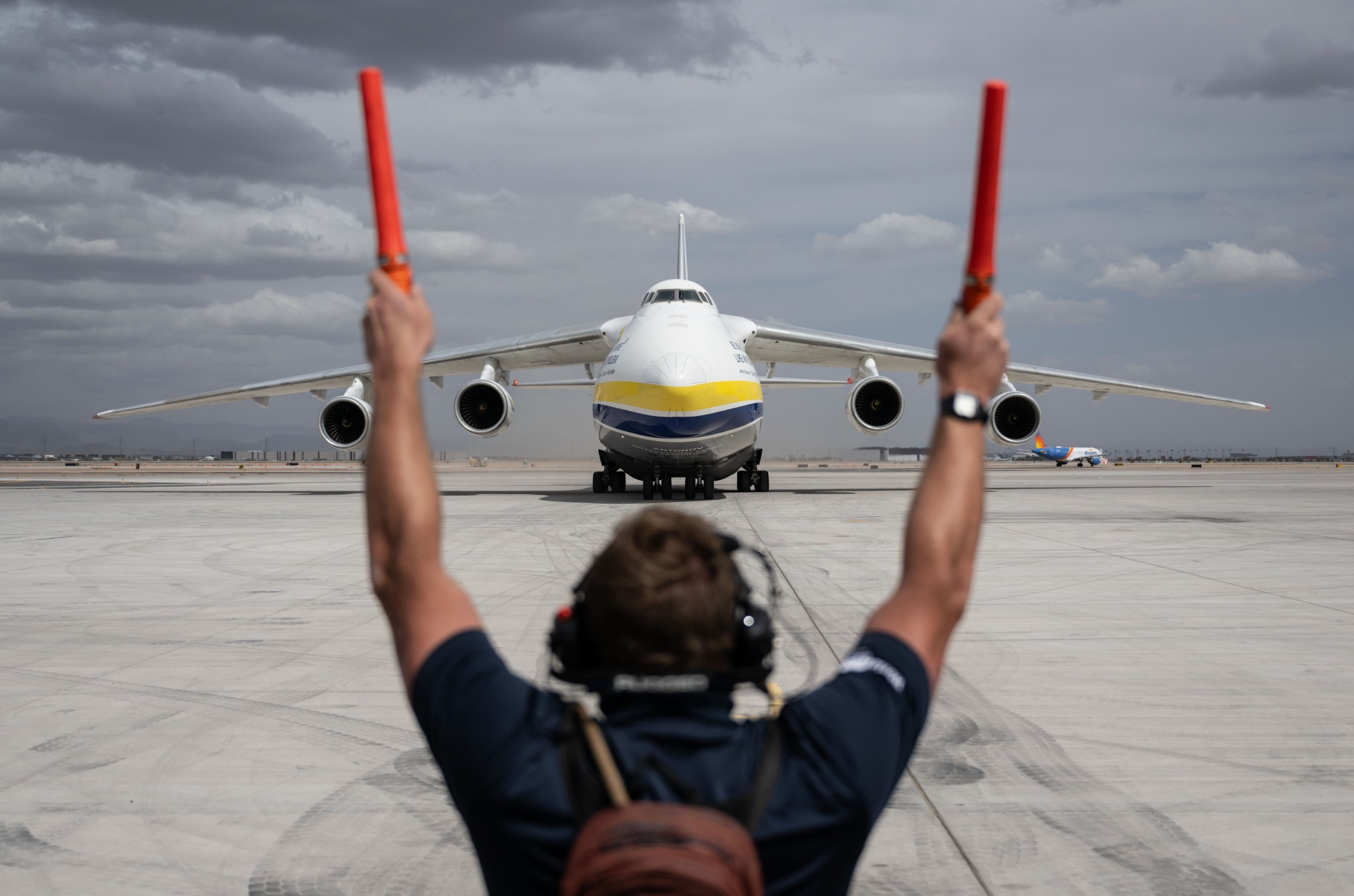 A ground crew member with arms raised and holding orange marshalling wands guides a large cargo aircraft as it taxis on the runway. The aircraft faces the camera head-on under a cloudy sky.