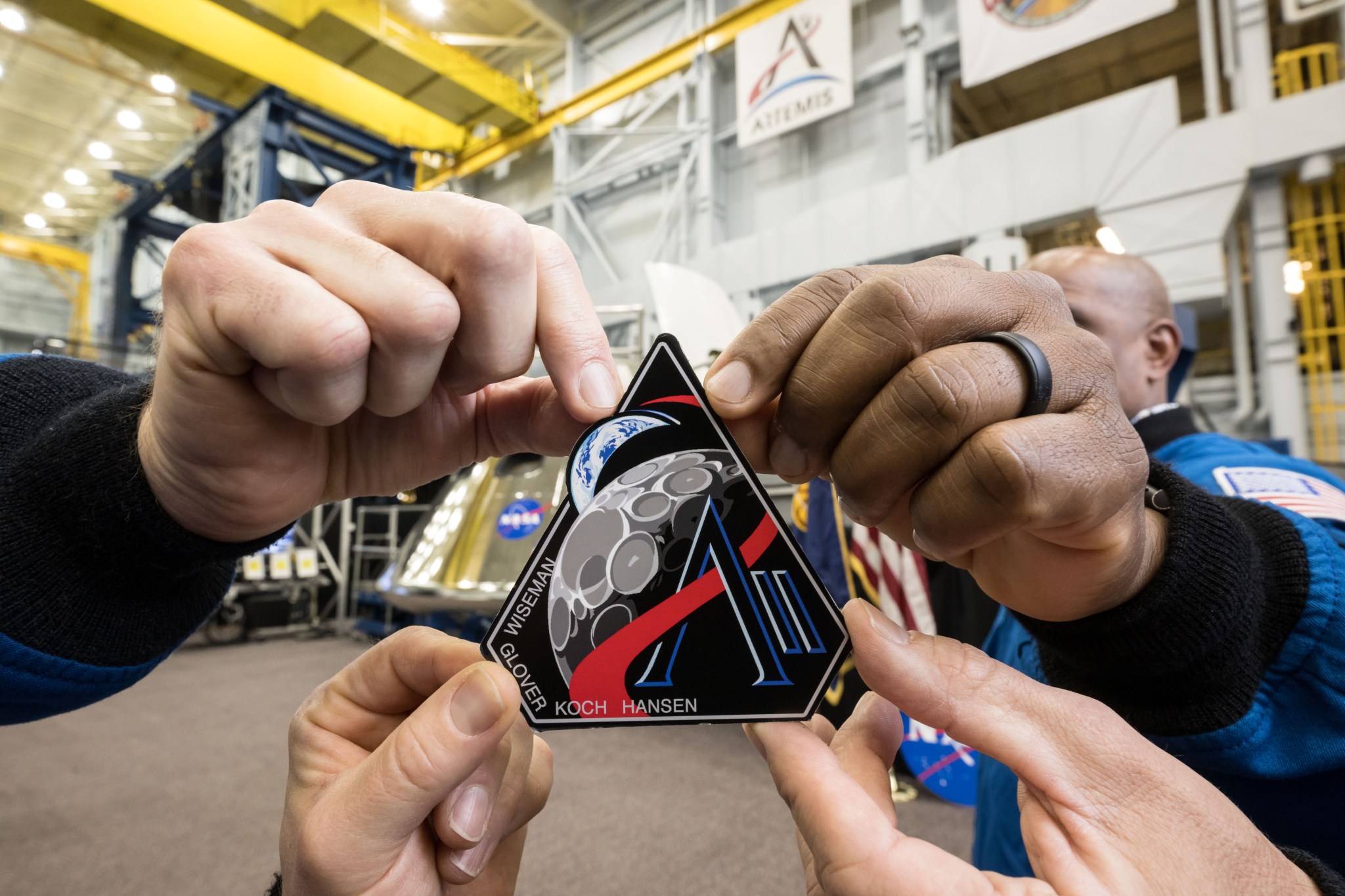 Four hands hold onto a corner of the Artemis II mission patch. The patch is five-sided, with a black background. The last names of the crew – Wiseman, Glover, Koch, and Hansen – are written along the patch’s bottom left corner. The Earth and Moon are visible on the patch, reminiscent of the famous “Earthrise” photo. A red line curves through the lines of a letter “A,” then around the Moon and Earth. Next to the “A” are two lines that look like the Roman numeral “I”: this represents Artemis II.